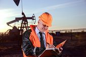 foto of sunset  - Oil worker in orange uniform and helmet on of background the pump jack and sunset sky - JPG 