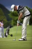image of golf  - Senior male golf player putting a golf ball on the green into the hole with female golf player waiting in the background - JPG 