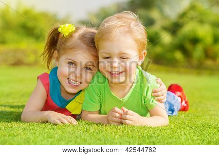 Picture or Photo of Image of two happy children having fun in the park, brother and sister lying down on green grass, best friends playing outdoors in spring, adorable little girl with cute boy enjoying springtime nature