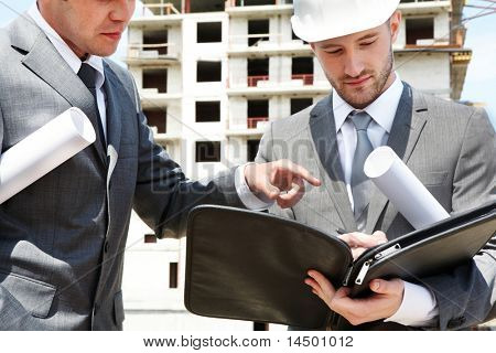 Picture or Photo of Portrait of two builders standing at building site and discussing new project held by one of them