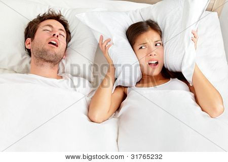 Picture or Photo of Snoring man. Couple in bed, man snoring and woman can not sleep, covering ears with pillow for snore noise. Young interracial couple, Asian woman, Caucasian man sleeping in bed at home.