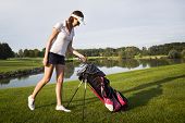 stock photo of golf  - Woman golf player taking out golf club from golf bag on golf course with beautiful pond and trees in background - JPG 