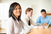 image of meeting  - An attractive Asian businesswoman looks at the camera during a meeting with a diverse group of business people including a latino and caucasian male - JPG 