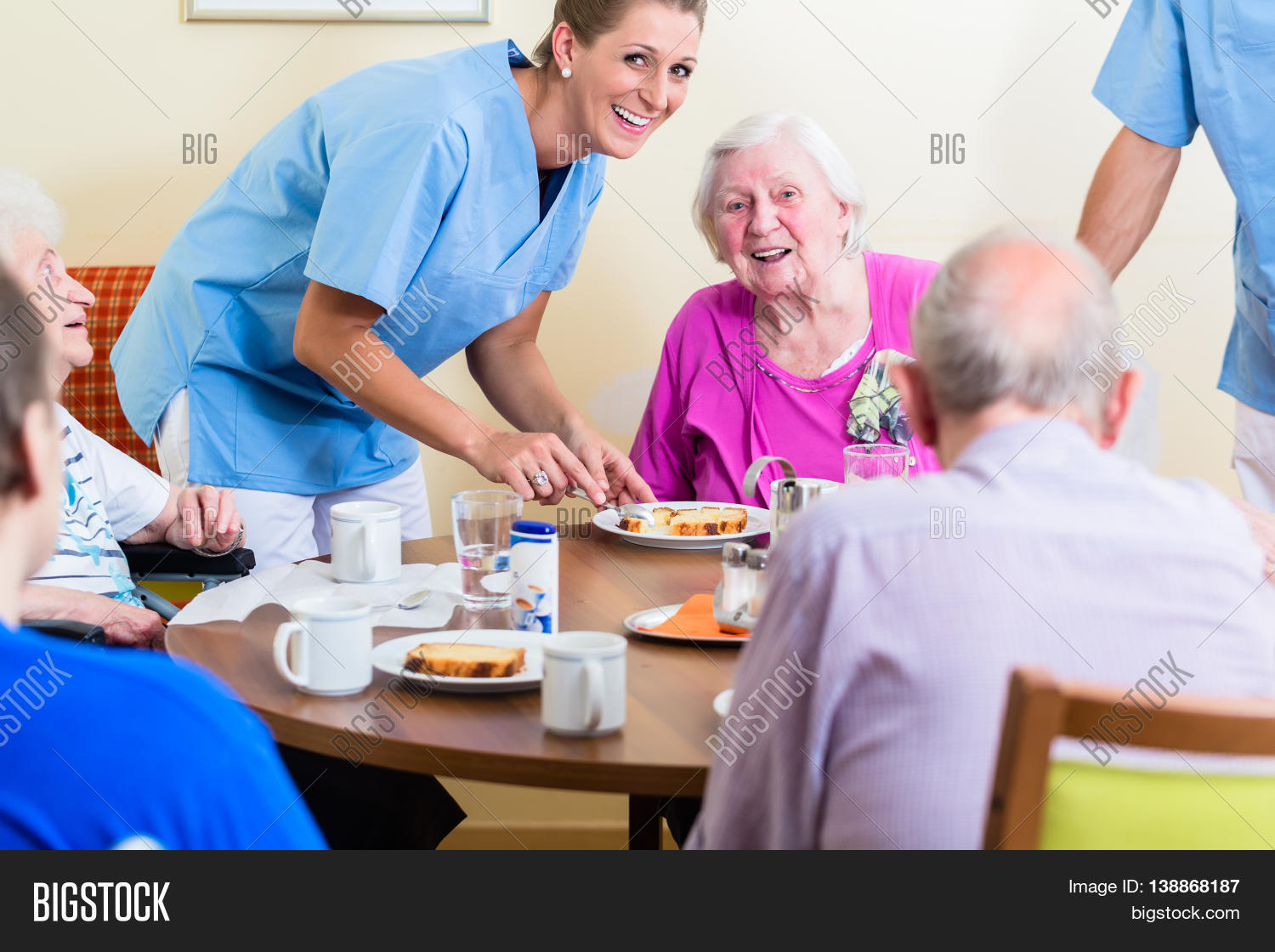 Group of seniors having food in nursing home, a nurse is serving Stock