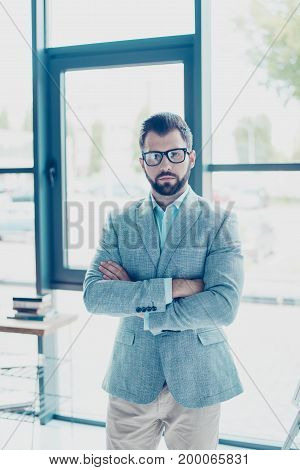 Young successful bearded nerdy man is standing in his office in formal suit and glasses with crossed hands looking serious severe harsh and strict. Behind him is a workplace and windows