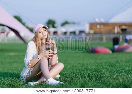 A stylish girl in a pink cap resting on a green lawn in the middle of a recreation park and holds a refreshing drink in her hands. Summer concept.