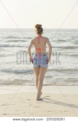 Young european female walking at the beach near seaside with waves. Young woman in pink swimsuit and denim shorts. Vacation and relax concept