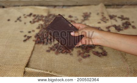 Woman hand holding a block of handmade chocolate with dry cocoa beans on a jute covered table in the background
