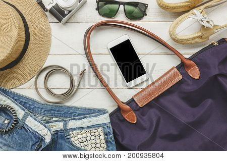 Top view accessories to travel with women clothing concept.white mobile phone belt bag hat camera necklace trousers and sunglasses on white wood table.