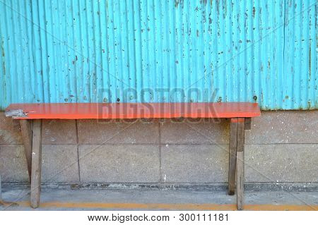 An Empty Old Orange Woiden Bench On The Ground Floor With A Brick Wall And Blue Zinc Wall