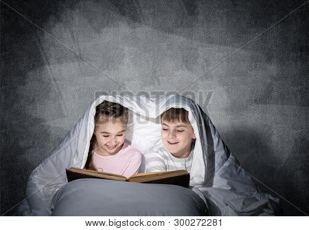 Engrossed Little Girl And Boy Reading Book In Bed After Bedtime. Pretty Kids Together Hiding Under B