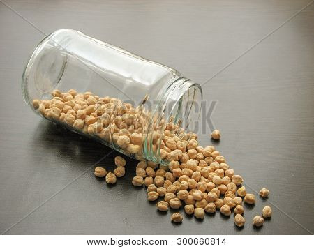 Dried Cheakpea Beans Are Poured From A Glass Jar On Brown Wooden Surface. Cheakpea Beans  In A Glass