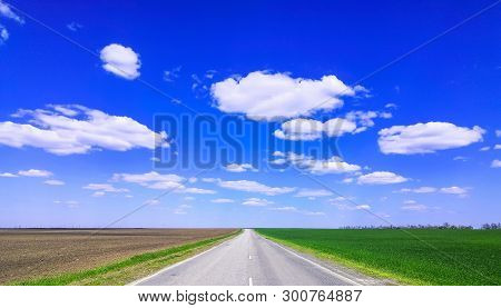 Empty Highway Through Green Grass Field Under White Clouds On Bright Blue Sky In Spring Summer Day. 