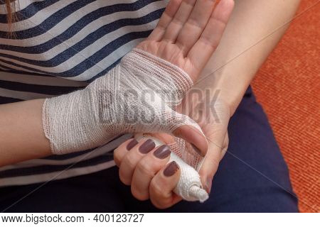 Young Woman Is Bandaging Her Wounded Palm And Wrist While Sitting On The Orange Sofa. View From Abov