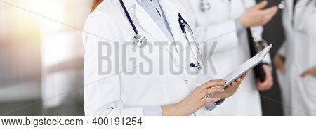 Unknown Woman Doctor Iusing Tablet Computer In Her Hands, While Standing In Sunny Clinic. Medicine C