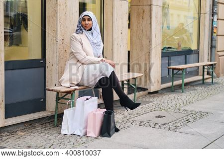 Young Beautiful Arabic Woman In Hijab Resting On Bench With Shopping Bags On Street