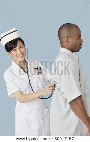 Portrait of a happy female doctor examining male patient's back with stethoscope over light blue background