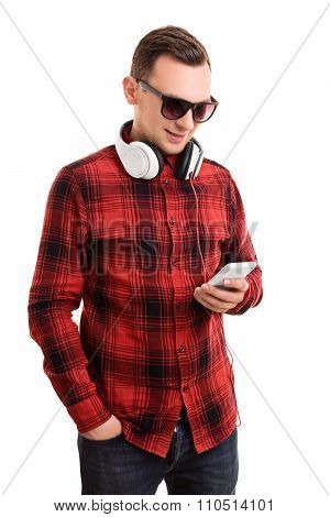 Modern and casual in style. Portrait of a confident smiling man or student in plaid shirt, glasses and headphones, looking at his mobile phone and texting, isolated on a white background.