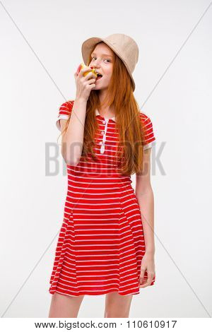 Amusing pretty cure redhead girl in boonie hat and red striped dress eating apple and posing on white background