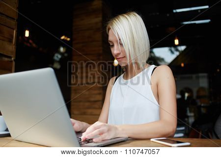 Young hipster girl keyboarding on her portable laptop computer while sitting in modern coffee shop