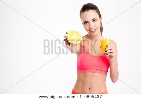 Positive attrative young sportswoman in pink top with glass of orange juice showing an orange half over white background