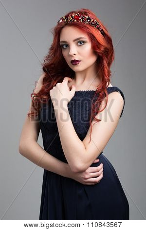 Beautiful red-haired fashion model posing in evening dress and in the diadem over dark background. f