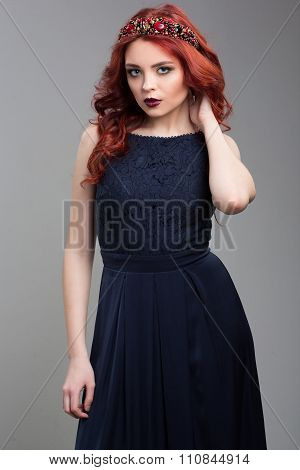 Beautiful red-haired fashion model posing in evening dress and in the diadem over dark background. f