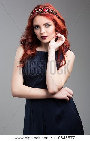 Beautiful red-haired fashion model posing in evening dress and in the diadem over dark background. f