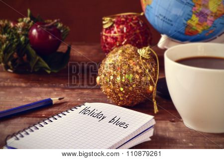 closeup of a notebook with the text holiday blues written in it on a rustic wooden table with some christmas ornaments, a cup of coffee and a globe
