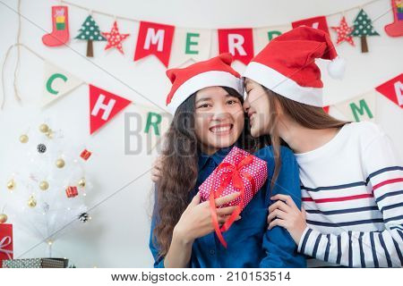 Asia lover Girlfriend kiss cheek and give Christmas gift at xmas partyAsia girl friends wear santa hat exchange red gift box with smiling facegift givingLovely lesbian couple