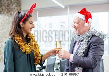 Happy colleagues talking at office party. Cheerful senior executive in Santa hat giving champagne flute to young female employee in antler headband. New Year in office concept