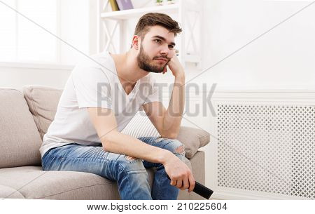 Young man watching television, using remote control to switch channels. Guy bored with what he sees on TV screen. Sitting on couch in living room at home, copy space