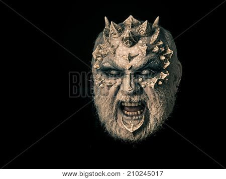 Evil face with dragon skin and grey beard. Man angry with blind eyes. Monster with thorns and horns. Demon baring teeth isolated on black. Horror and fantasy concept.