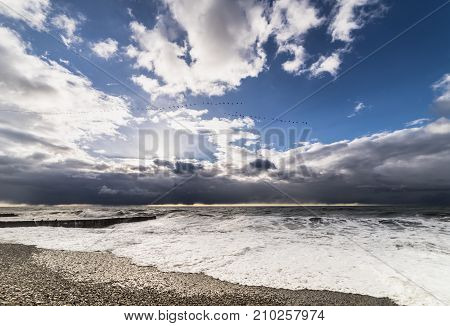 The raging waves are washed by a stone beach against a blue sky with white clouds