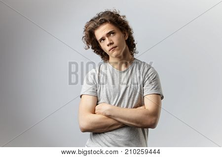 Studio shot of pensive young European male with voluminous hair posing at blank wall keeping arms crossed and looking up with thoughtful expression making plans for weekend. Body language