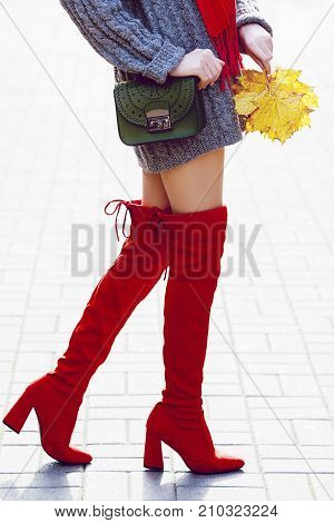Fashionable woman wearing trendy red high, over the knee suede boots walking in street. Model holding small green textured bag and yellow autumn leafs. Elegant city outfit. Female fashion concept