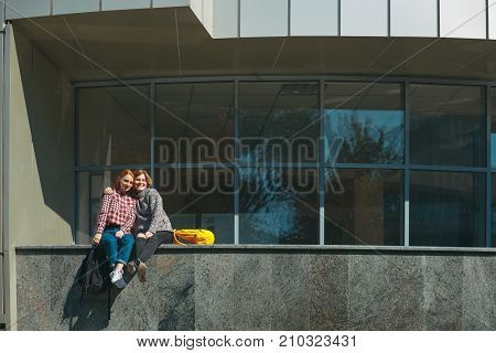 College friends take a break after classes embracing each other. Young girls with reddish hair sit on a modern building near windows on campus outdoors. Friendship and studentship.
