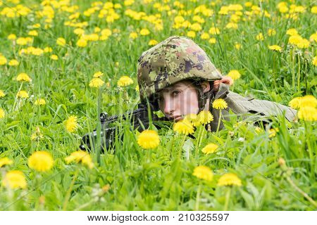 Portrait of armed woman with camouflage. Young female soldier observe with firearm. Child soldier with gun in war green goutweed and yellow dandelions background. Military army people concept