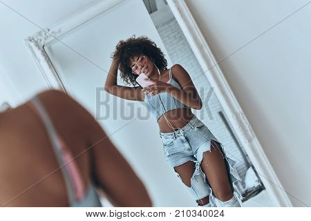 Girl with a great style. Reflection of beautiful young African woman listening music and keeping hand in hair while taking selfie in the mirror at home