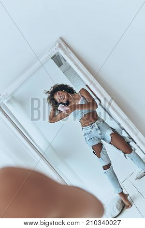 Perfect selfie. Reflection of beautiful young African woman listening music and keeping hand in hair while taking selfie in the mirror at home