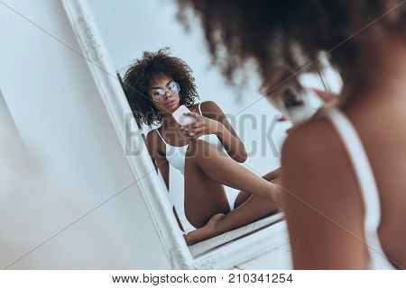 Her body is perfection. Reflection of beautiful young African woman in white lingerie taking selfie in the mirror while sitting on the floor at home
