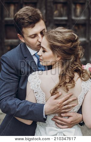 Happy Couple Of Newlyweds Posing Near Ancient Wooden Castle Gates, Handsome Smiling Groom Embracing