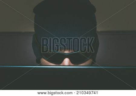 Hacker hacking confidential information on his computer while he is wearing hat and sunglasses close up