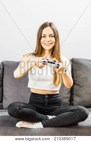 Pretty teenager girl playing a video game joyfully on the comfortable grey sofa before white wall