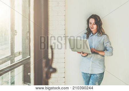 Front view.Young woman dressed in blue shirt is standing in room near white wall and is using laptop.Hipster girl checks email on computer, blogging, chatting, shopping online.Online marketing, education.