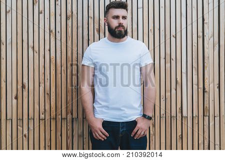 Summer day. Front view. Young bearded hipster man dressed in white t-shirt is stands outdoor against wood wall. Mock up. Space for logo, text, image.