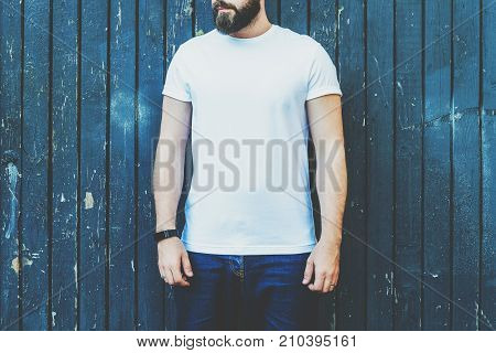 Summer day. Front view. Young bearded hipster man dressed in white t-shirt and sunglasses is stands against dark wood wall. Mock up. Space for logo, text, image.