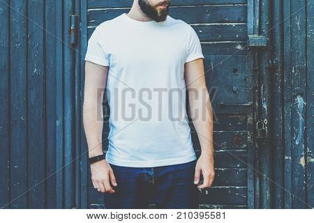 Summer day. Front view. Young bearded hipster man dressed in white t-shirt and sunglasses is stands against dark wood wall. Mock up. Space for logo, text, image. Advertising.