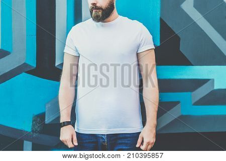 Summer day. Front view. Young bearded hipster man dressed in white t-shirt is stands outdoor against wall with graffiti. Mock up. Space for logo, text, image.