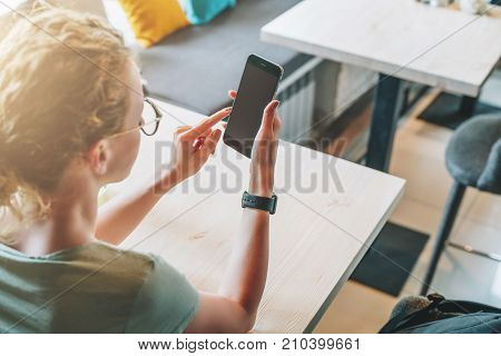 Rear view. Hipster girl is sitting in cafe at table, using digital gadget, looking on screen of smartphone, checking email. Businesswoman working online.